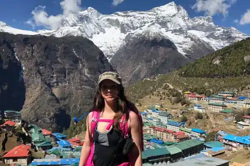 A female traveler with a view towards Namche in the background, where trekkers are acclimatizing to make their Everest Base Camp Trek successful .